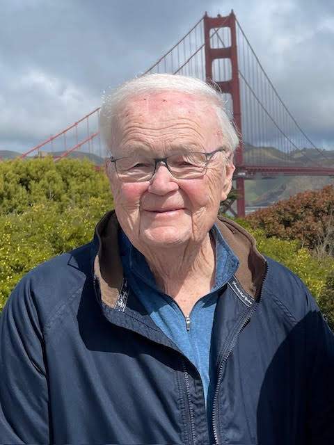 An elderly man with white hair and glasses smiles in front of the Golden Gate Bridge on a partly cloudy day, wearing a blue jacket and denim shirt.