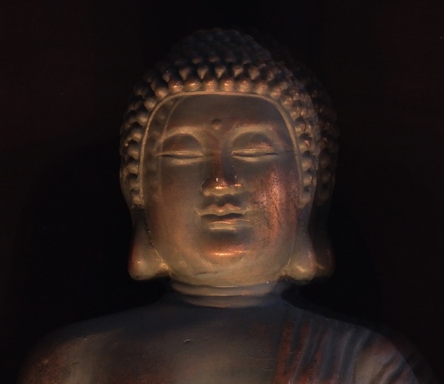 A dimly lit budha statue with closed eyes and a serene expression, featuring curled hair and detailed facial features, stands gracefully against a dark background.