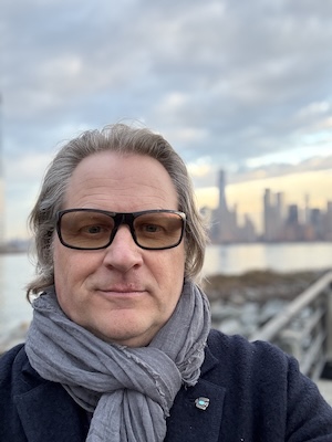 A person wearing dark-rimmed glasses, a gray scarf, and a dark coat stands outdoors near water with a city skyline in the background under a cloudy sky, embodying the calm of Infinite Circle Zen.