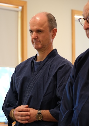 A man in a navy blue martial arts uniform stands indoors with his hands clasped, looking focused—embodying the calm presence of Infinite Circle Zen. Another person in similar attire is partially visible beside him.