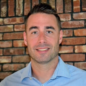 A man with short brown hair and stubble, wearing a light blue checkered shirt, smiles in front of a brick wall background, evoking the calm and balance of Infinite Circle Zen.