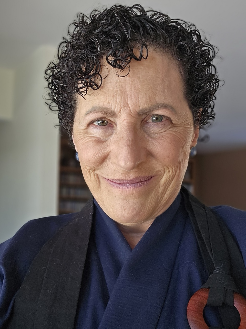A person with short, curly dark hair and light skin smiles gently at the camera. Wearing a navy blue robe, black scarf, and a wooden ornament, they embody the calm presence of Infinite Circle Zen against a softly blurred background.