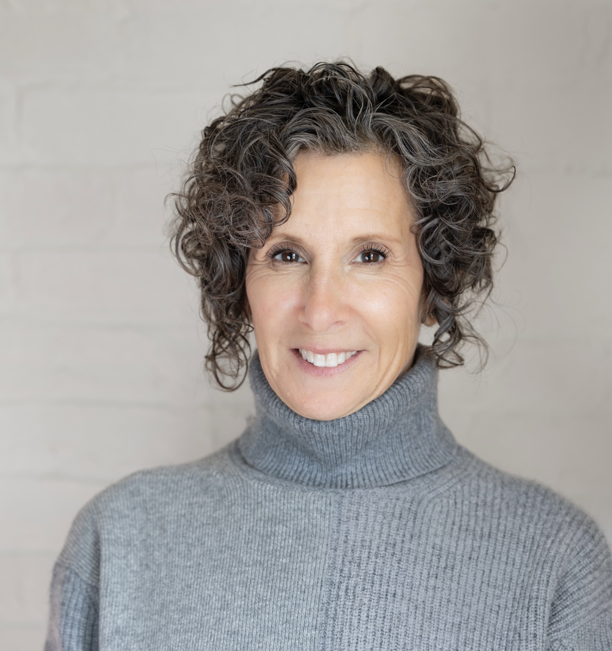 A woman with short, curly gray hair smiles warmly. She is wearing a gray turtleneck sweater and stands in front of a light-colored, textured wall, evoking the calm presence of Infinite Circle Zen.