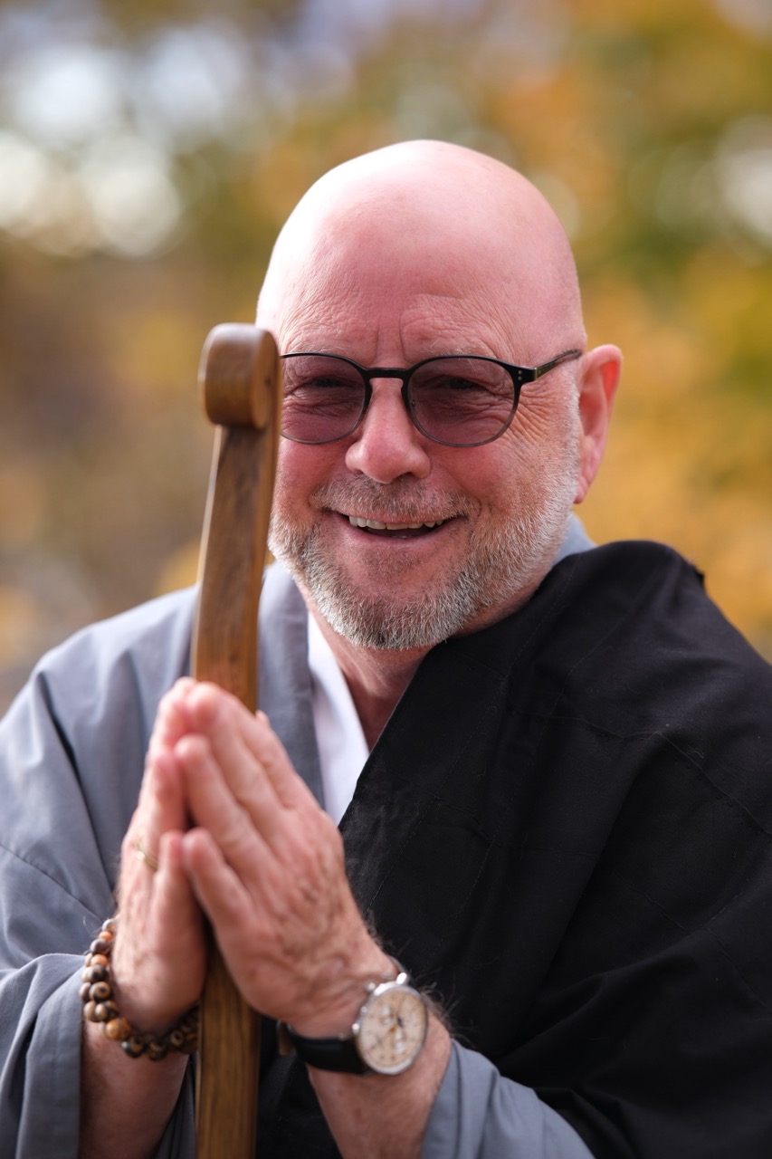 A smiling older man with glasses and a bald head holds a wooden staff, hands clasped in an Infinite Circle Zen gesture. He wears a beaded bracelet, wristwatch, and robe, with autumn foliage softly blurred in the background.