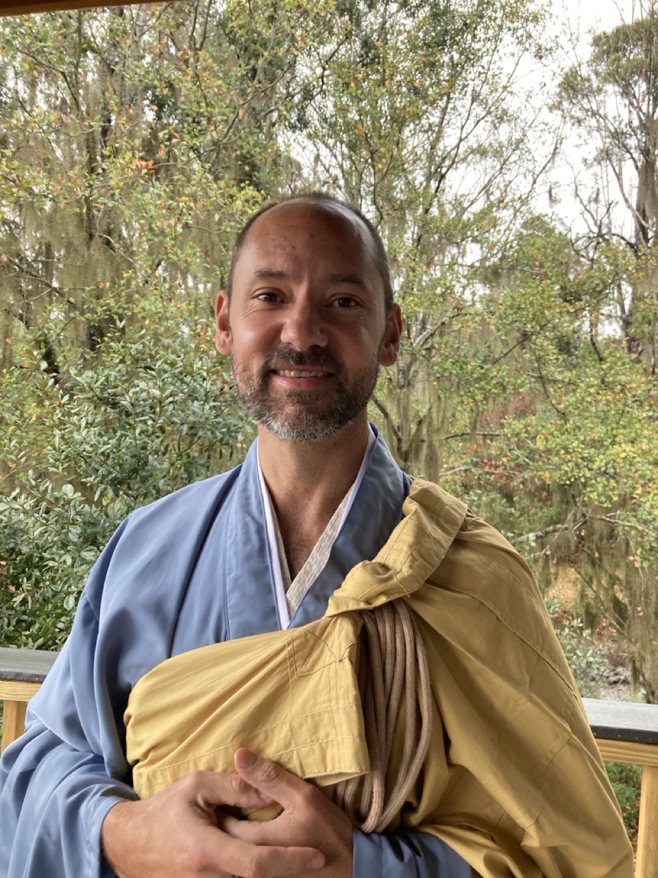 A man with a trimmed beard, wearing blue and yellow Buddhist robes, stands on a wooden deck with green trees in the background, smiling gently at the camera—embodying the calm presence of Infinite Circle Zen.