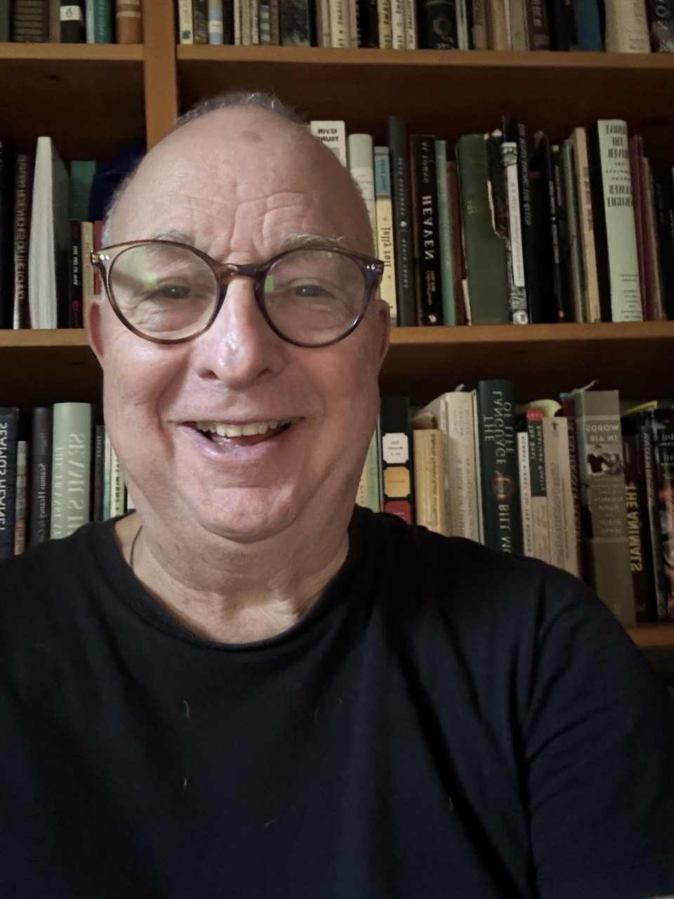 A smiling older man with glasses and a black shirt takes a selfie in front of a bookshelf filled with various books, including titles on Infinite Circle Zen.