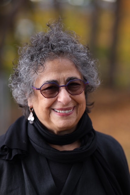 An older woman with curly gray hair, wearing purple glasses, earrings, and a black scarf smiles outdoors with autumn colors blurred behind her, radiating the calm presence of Infinite Circle Zen.