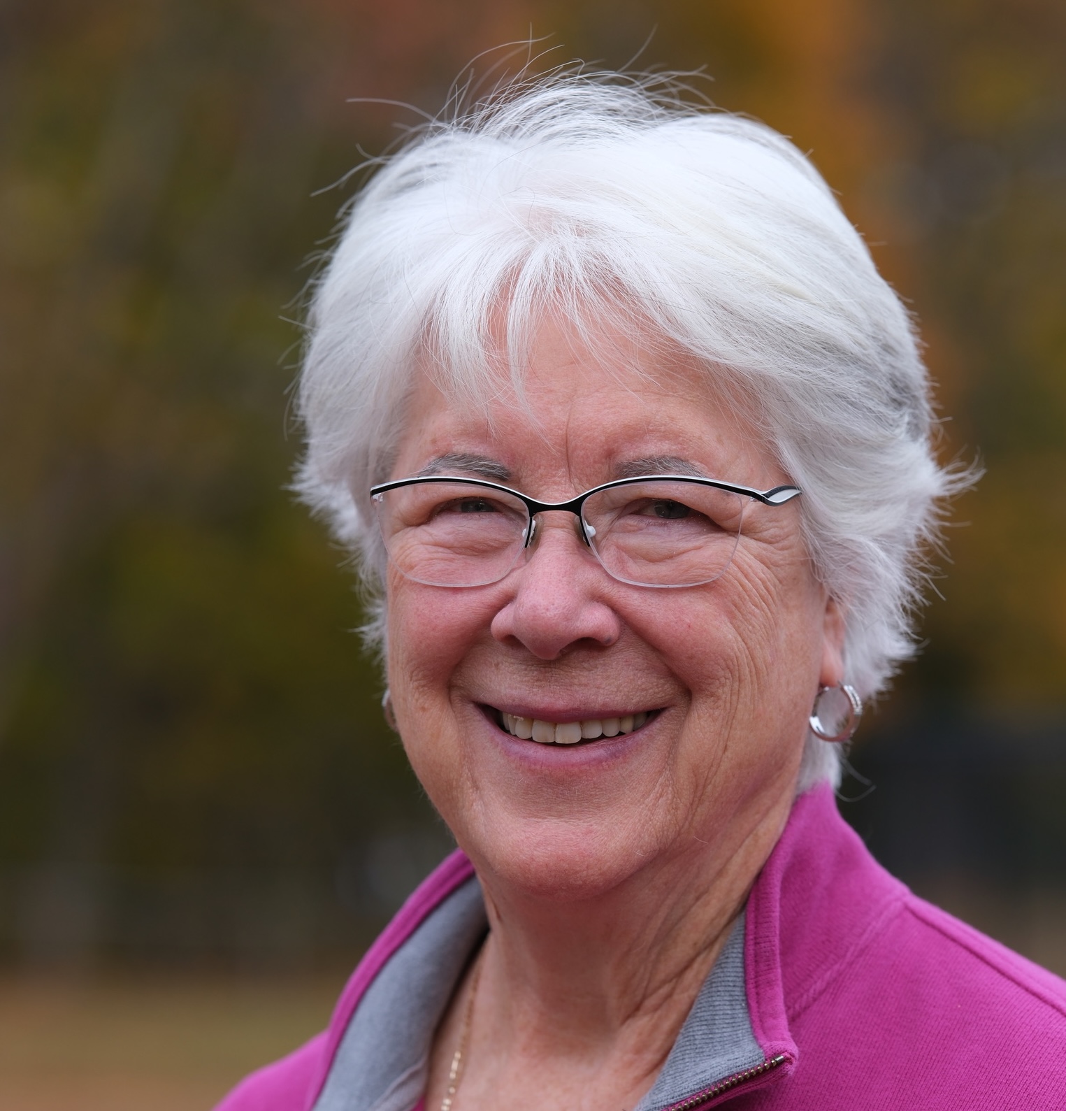 An older woman with short white hair and glasses smiles outdoors. She is wearing a pink jacket and silver hoop earrings, with a blurred background of autumn trees.