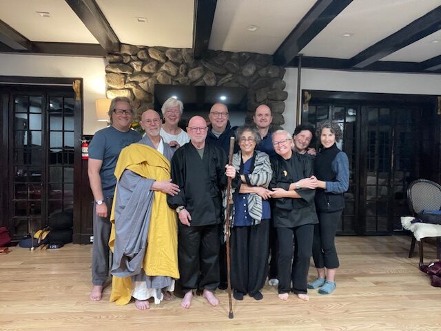 A group of ten people, some in traditional robes, stand closely together smiling in a warmly lit room with wooden floors, stonework, and beams. One person in front holds a wooden staff.