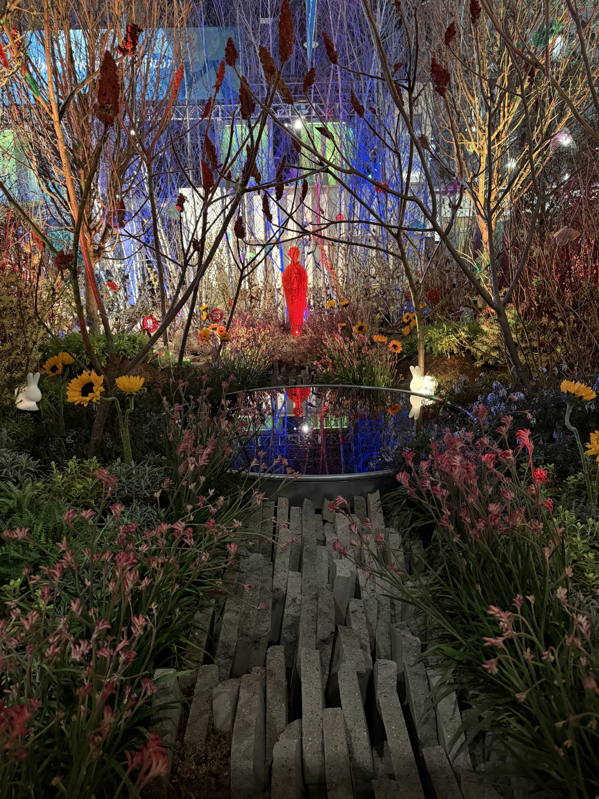 A vibrant garden with colorful flowers, tall branches, and a small pond. A person in a bright red outfit stands at the center, surrounded by dramatic lighting and reflections in the water.