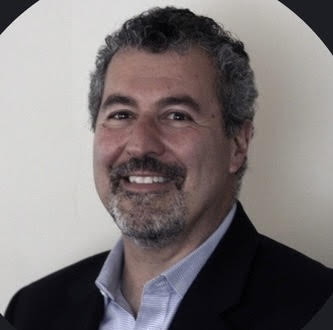 A man with short curly gray hair, a beard, and mustache is smiling. He is wearing a dark suit jacket over a light-colored collared shirt, standing in front of a plain light background.